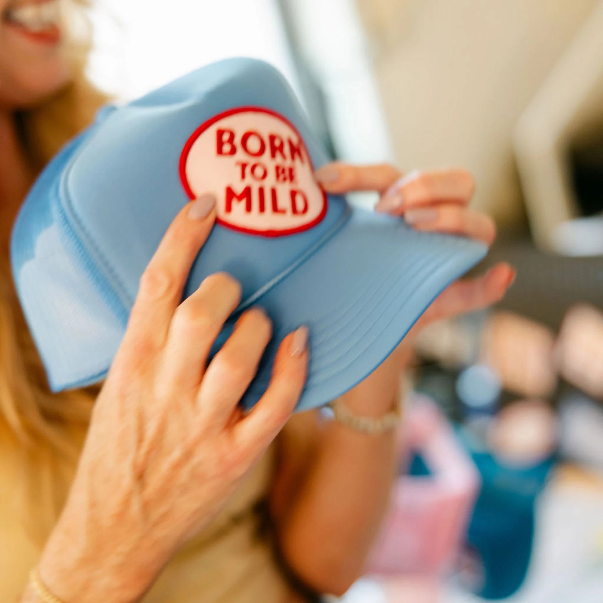 Woman holding a Born To Be Mild trucker hat in cobalt blue, showing the embroidered patch up close