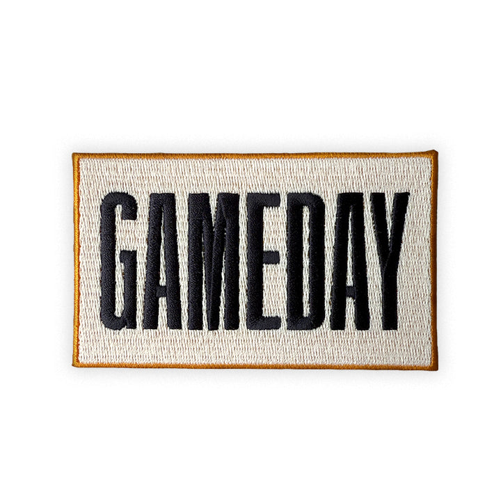 Gameday embroidered iron-on patch in cream with an antique gold border and smoky charcoal lettering on a plain white background